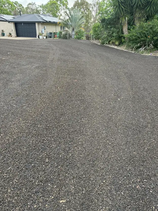 A wide bitumen driveway with a coarse grey stone seal surface leading up to a single-storey brick home with a dark garage door, bordered by garden and trees