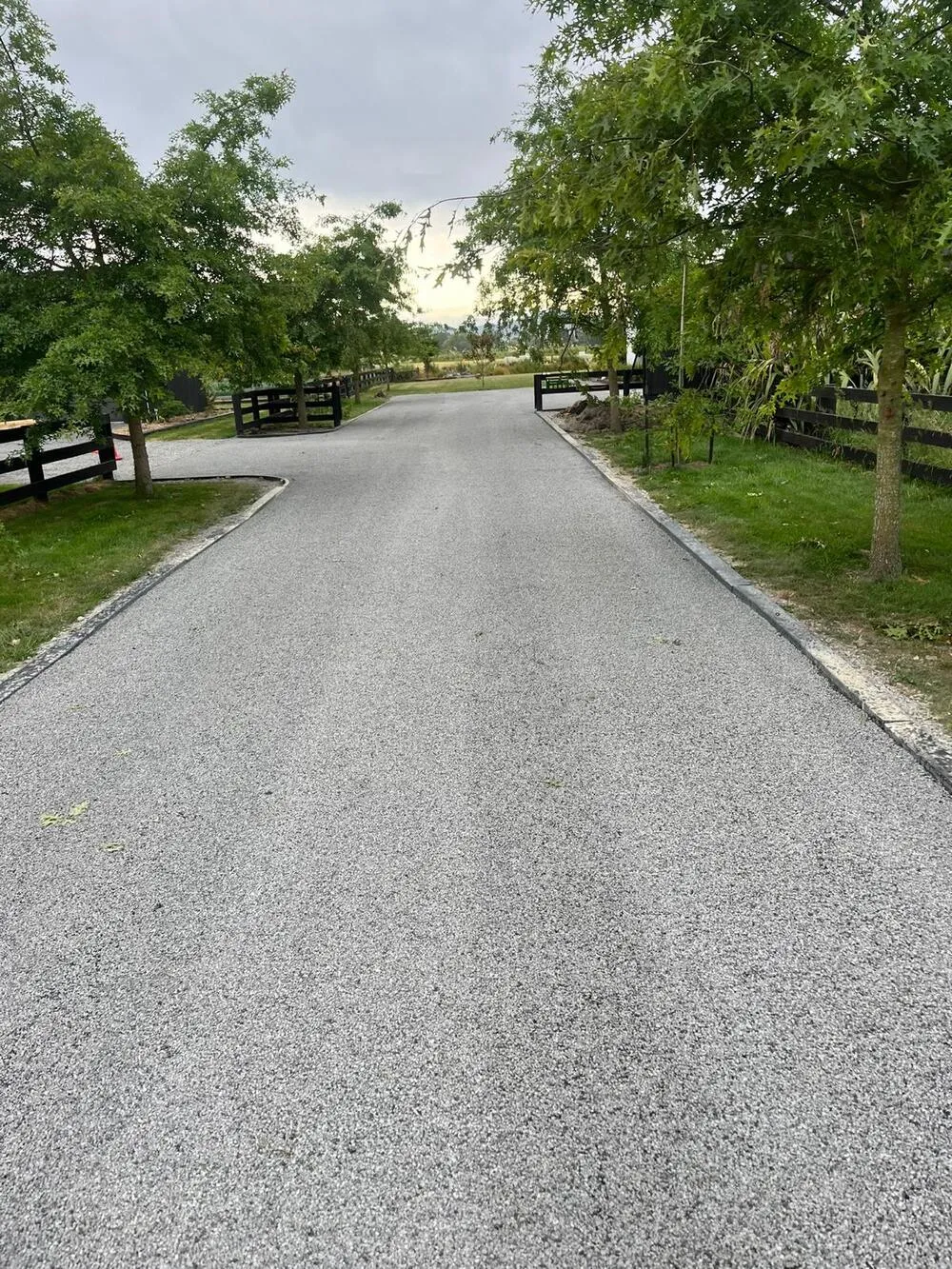 A sealed tree-lined driveway leading to a rural property entrance with black timber gates