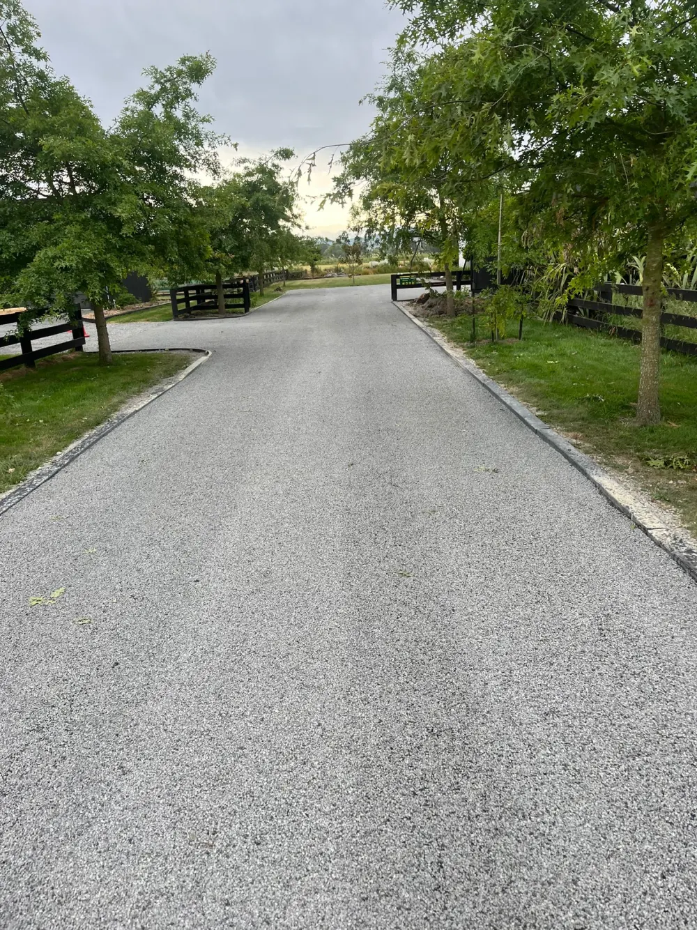 A long driveway with a light grey chip-seal aggregate surface, lined with black timber post-and-rail fencing, trees and lawn under an overcast sky
