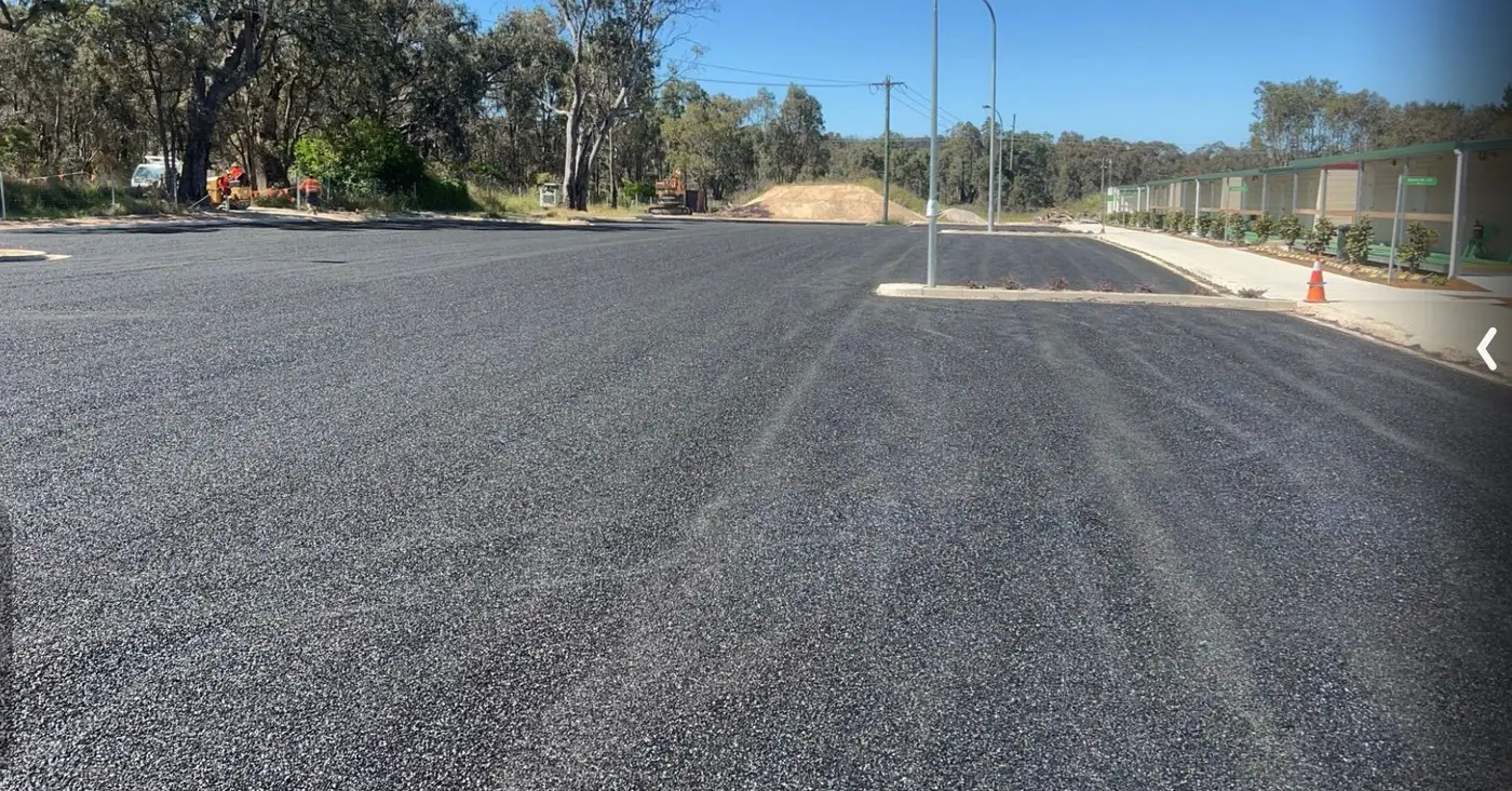 A wide expanse of freshly laid asphalt at ground level, with a light pole, traffic cone and gum trees along the edge under a clear blue sky