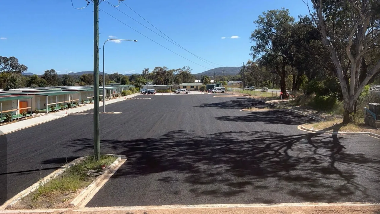 A freshly laid asphalt car park bordered by new kerbing and garden beds, with a single-storey building, gum trees and bushland hills under a clear blue sky