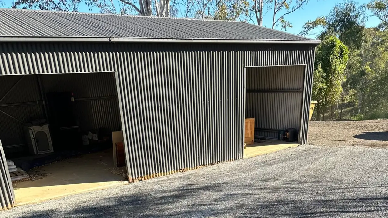 A bitumen spray seal driveway of dark stone aggregate in front of a large steel shed on a rural property, with gum trees and blue sky behind