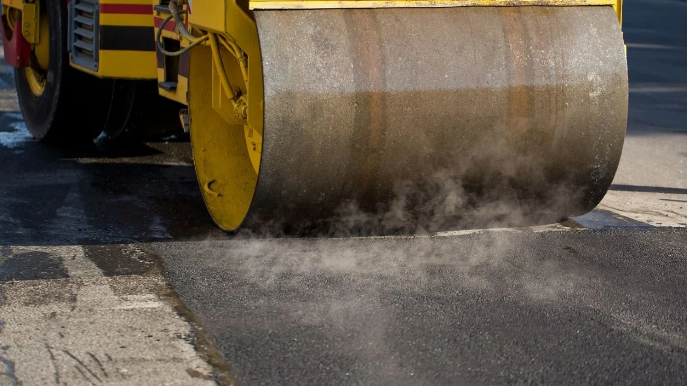 A steel drum roller compacting fresh hot mix asphalt, steam rising off the surface